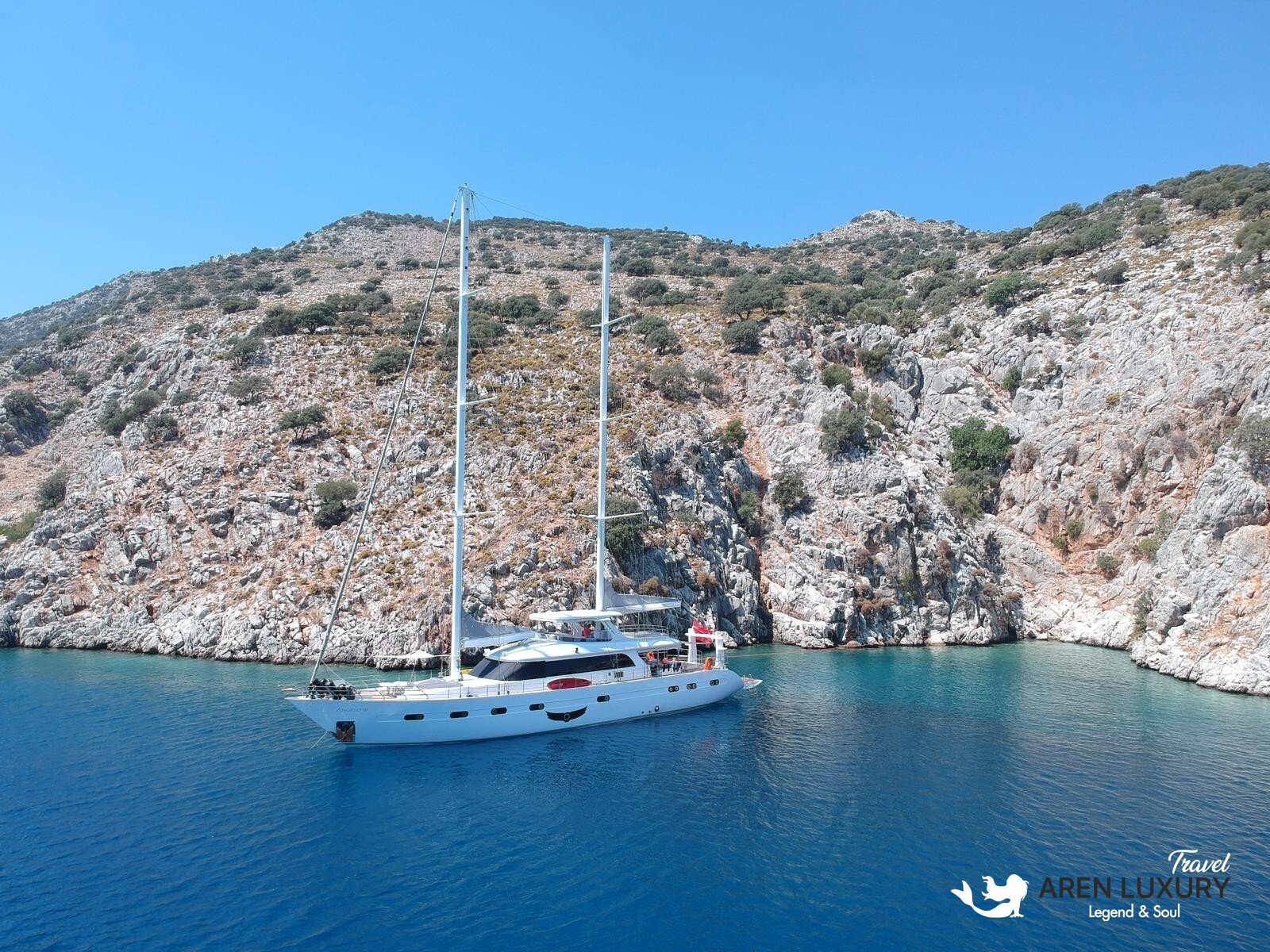 High-angle aerial view of the 35m Angelo 3 motorsailer yacht anchored in a deep blue Mediterranean bay.