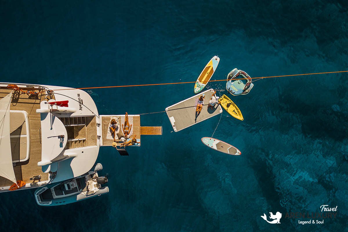 Aerial top-down view of the 35m Angelo 3 motorsailer with guests enjoying a swim platform, paddleboards, and water toys in deep blue water.