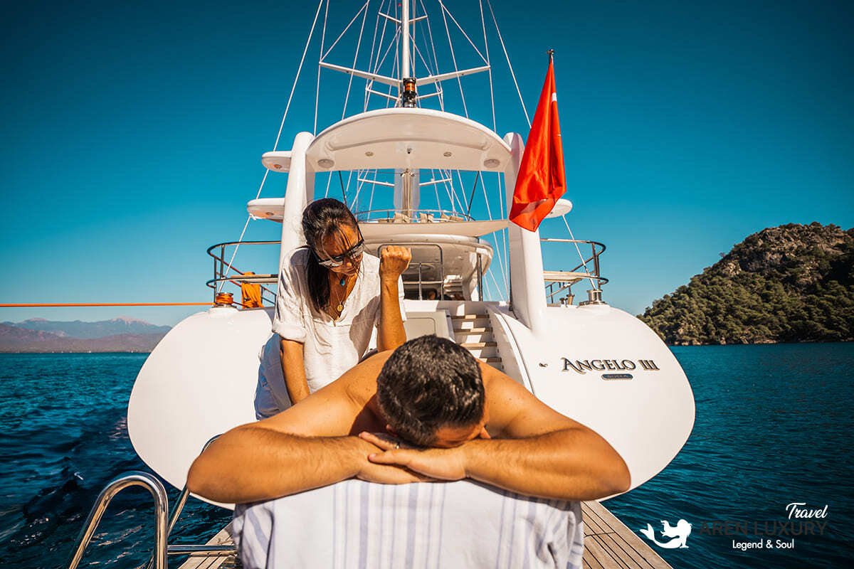 Side profile of the Angelo 3 motorsailer showing the sleek hull, flybridge, and dual masts against a coastal backdrop