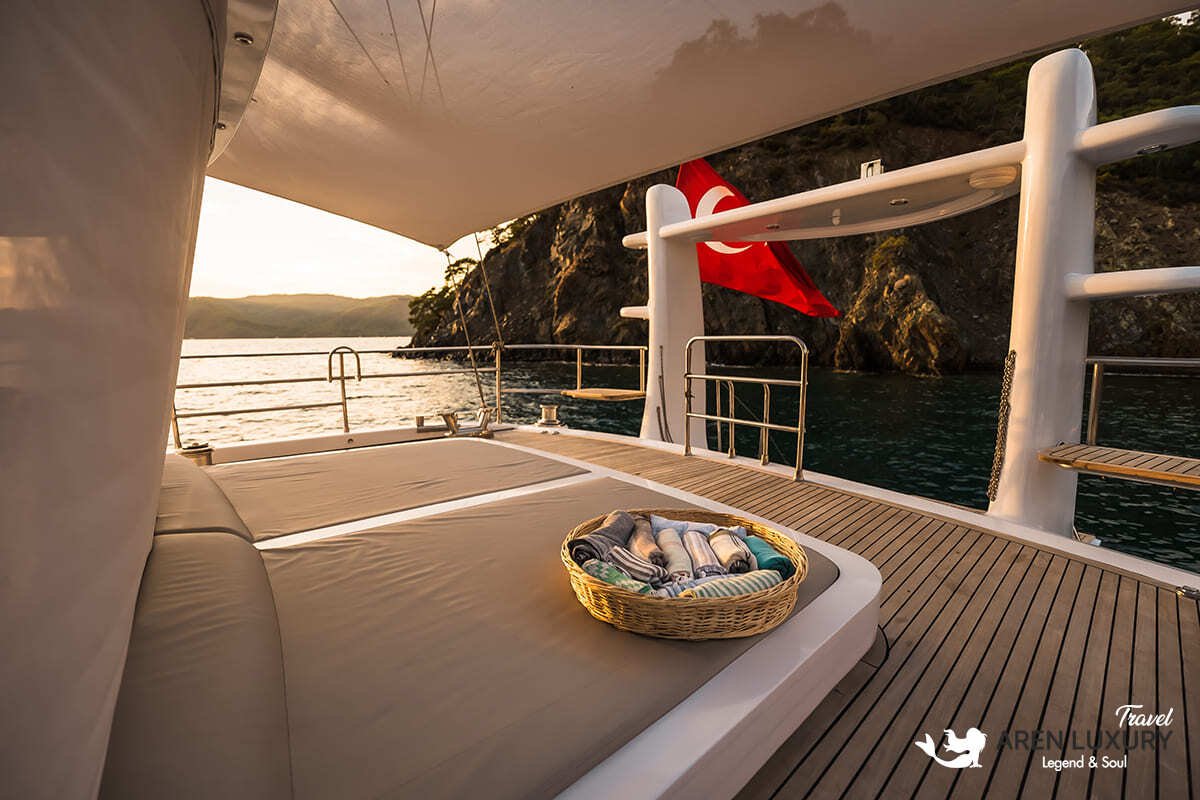 Side profile of the Angelo 3 motorsailer showing the sleek hull, flybridge, and dual masts against a coastal backdrop