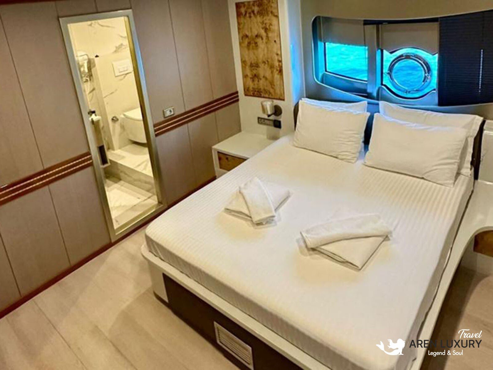 Modern luxury cabin interior of Angelo 3 motorsailer with a large bed, en-suite bathroom, and circular porthole window.