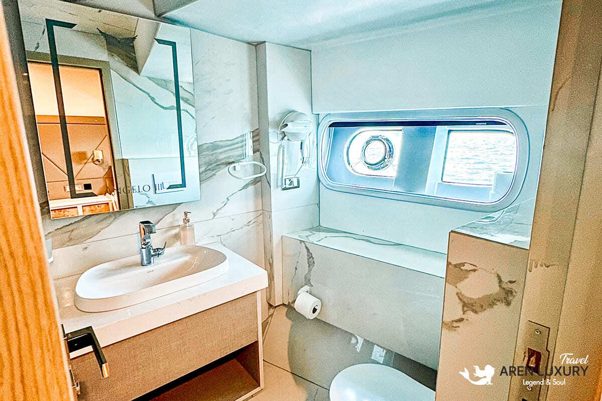 Modern luxury cabin interior of Angelo 3 motorsailer with a large bed, en-suite bathroom, and circular porthole window.