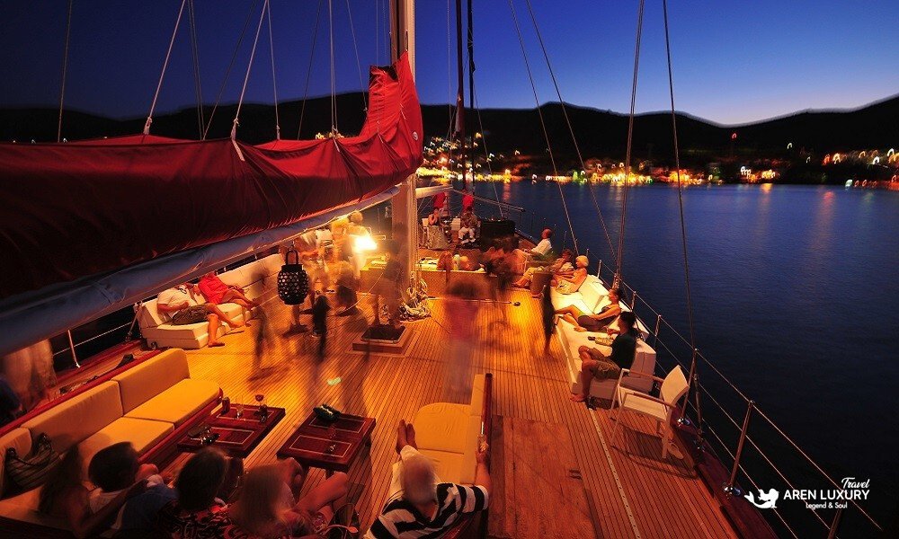 Casa-Dell-Arte-2-Gulet-Night-Event-Bodrum Vibrant evening social gathering on the illuminated teak deck of Casa Dell'Arte 2 luxury gulet at anchor in a Bodrum bay.