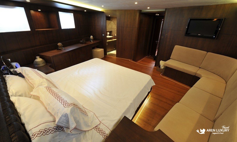 Casa-Dell-Arte-2-Gulet-Master-Suite-Interior-Amenities Wide-angle view of the master suite on Casa Dell'Arte 2 luxury gulet, featuring a queen-sized bed, integrated L-shaped sofa seating, mahogany cabinetry, and a flat-screen TV.