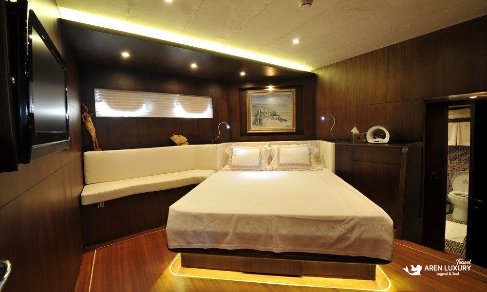 Casa-Dell-Arte-2-Gulet-Luxury-Double-Cabin-Interior Luxurious double guest cabin on the Casa Dell'Arte 2 gulet featuring a queen bed with ambient LED lighting, mahogany wood paneling, contemporary art, and a view into the private ensuite bathroom.