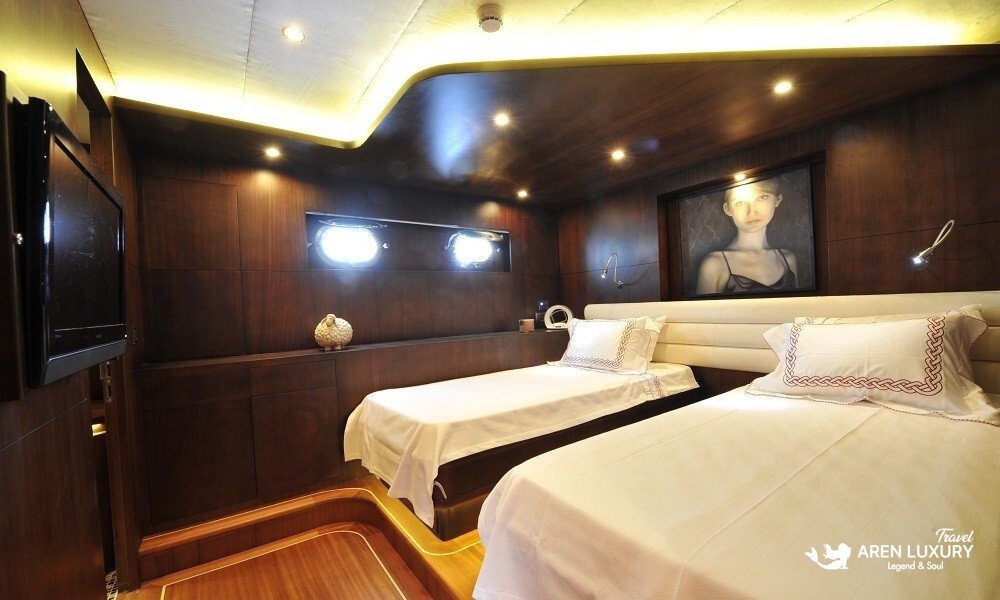 Casa-Dell-Arte-2-Gulet-Twin-Guest-Cabin Elegant twin guest cabin on Casa Dell'Arte 2 luxury gulet, featuring two single beds, contemporary wall art, polished mahogany walls, and ambient LED lighting.