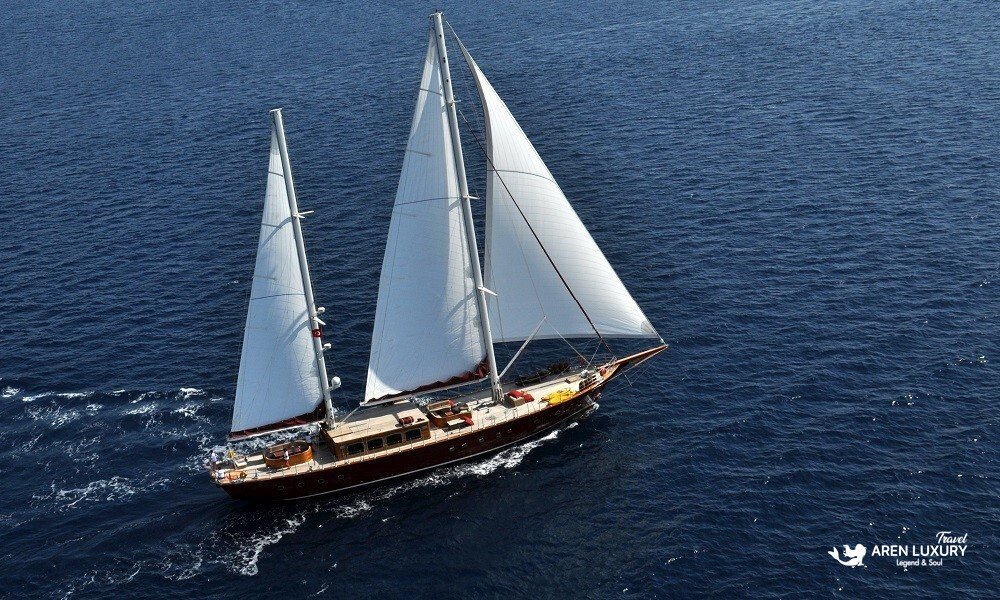 Casa-Dell-Arte-2-Luxury-Gulet-Aerial-Sailing-Bodrum High-angle aerial view of the Casa Dell'Arte 2 luxury gulet sailing the deep blue waters of Bodrum, showcasing its full white sails and elegant wooden design.
