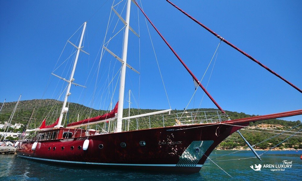 Casa-Dell-Arte-2-Luxury-Gulet-Side-Profile-Bodrum Side profile of the Casa Dell'Arte 2 luxury gulet docked in a crystal-clear bay in Bodrum, Turkey, showcasing its majestic mahogany hull and tall masts against a blue sky.