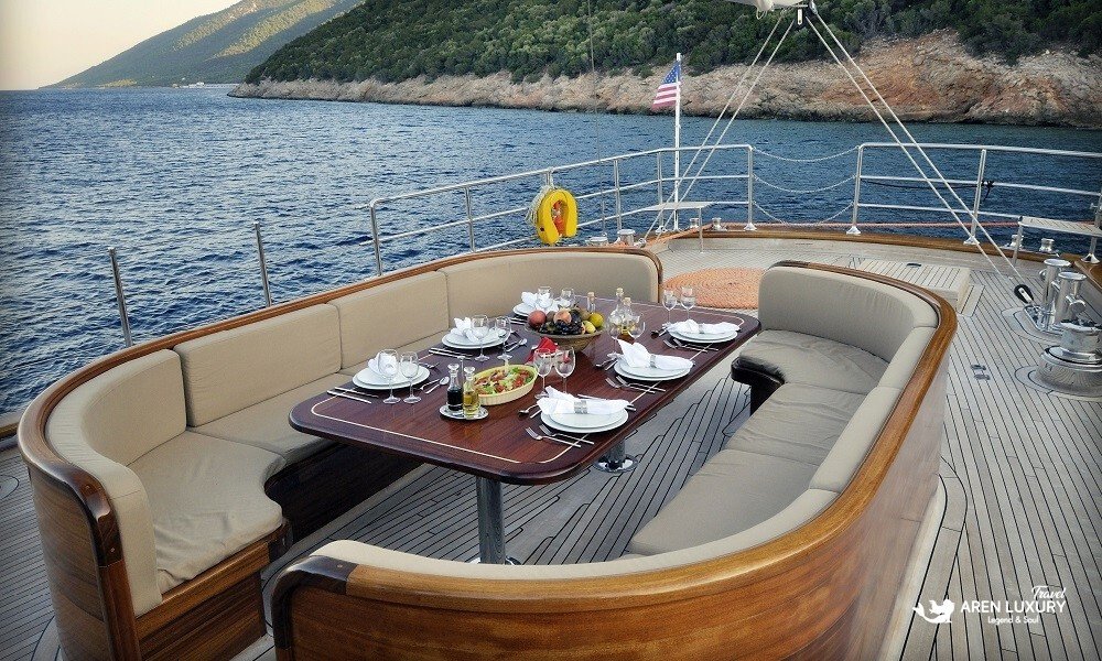 Casa-Dell-Arte-2-Gulet-Alfresco-Dining-Area Elegant alfresco dining table on the teak deck of Casa Dell'Arte 2 luxury gulet, set for a gourmet meal with a scenic Mediterranean coastline background.