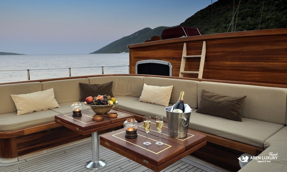 Luxury-Gulet-Sunset-Aft-Deck-Lounge-Bodrum Luxurious aft deck lounge of a high-end gulet at sunset in Bodrum, featuring champagne service, plush grey outdoor sofas, and a teak wood deck with scenic Mediterranean views.