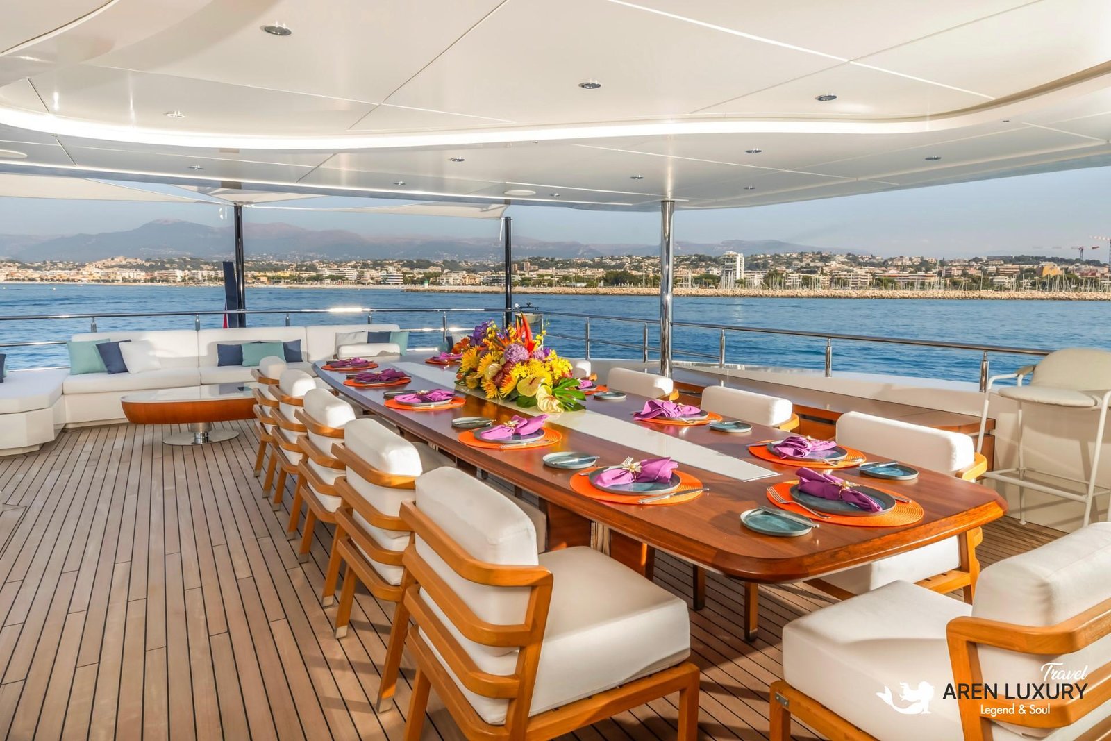 Formal long dining table set on the teak aft deck of Eternal Spark yacht with purple napkins and floral centerpieces against a coastal backdrop.
