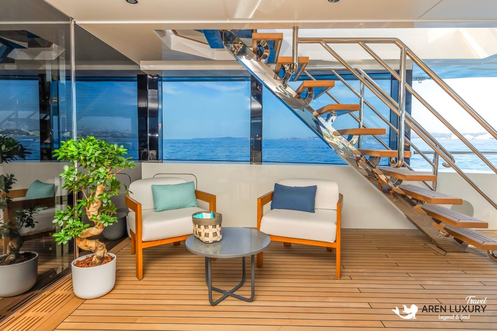Modern lower deck lounge area on the Eternal Spark yacht featuring white armchairs, a bonsai tree, and a sleek teak and stainless steel staircase.