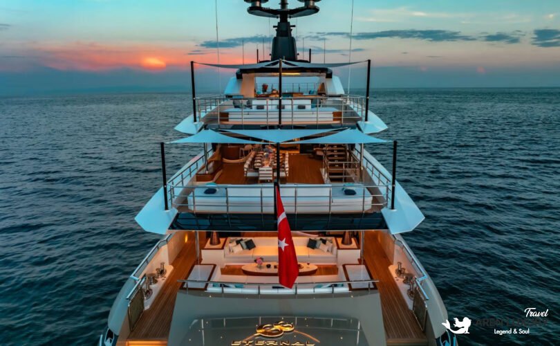 Rear view of the Eternal Spark yacht at sunset, showcasing multiple deck levels, outdoor dining areas, and warm interior lighting.