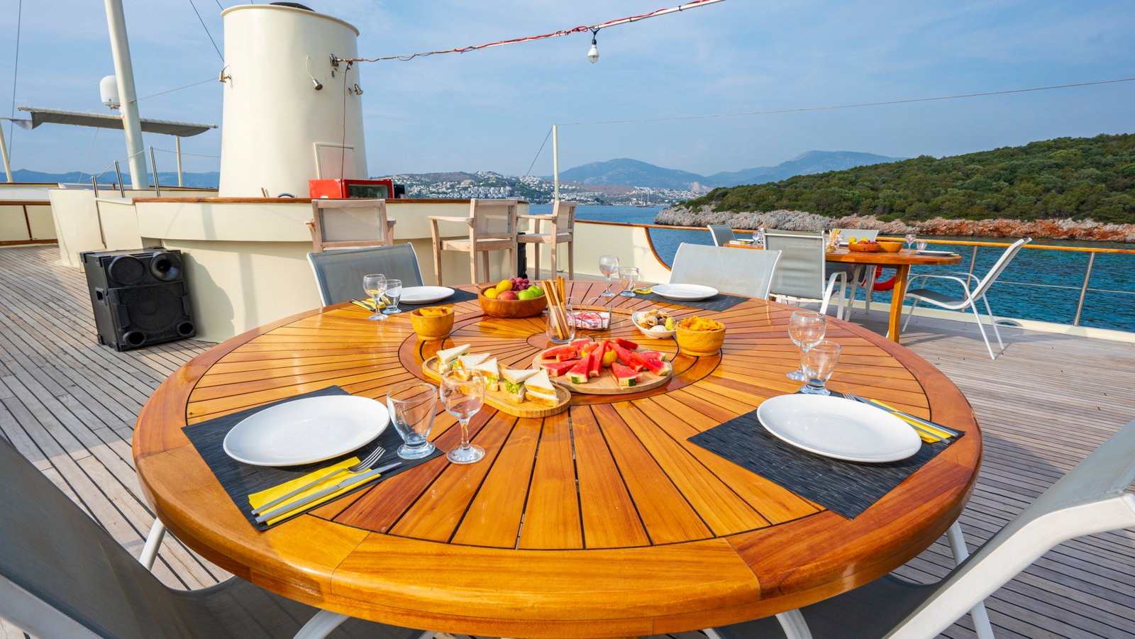 elara-1-gulet-outdoor-dining-deck Close-up of a large round wooden dining table on the deck of Elara 1 gulet with fresh fruit and appetizers.