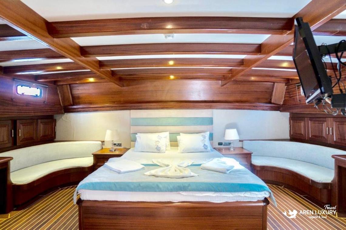 gulet-freedom-luxury-master-suite-interior-bodrum Luxury master cabin on Gulet Freedom featuring a large double bed with light blue accents, curved white lounge seating, and a flat-screen TV.