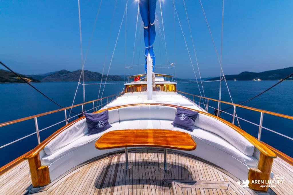 Luxury forward deck lounge on Gulet Gokce at sunset with curved seating, a polished wooden table, and blue sails overhead.