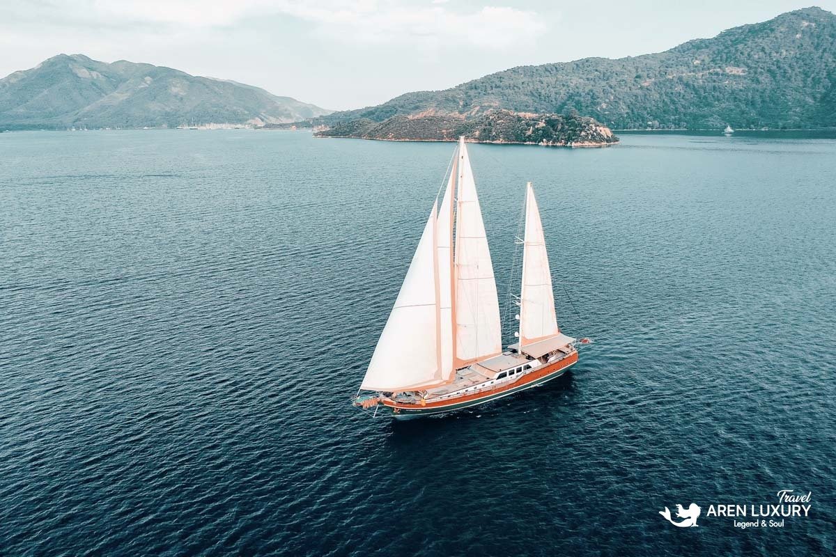 Wide aerial shot of the La Bella Vita gulet sailing with full white sails in a calm bay near green mountains.