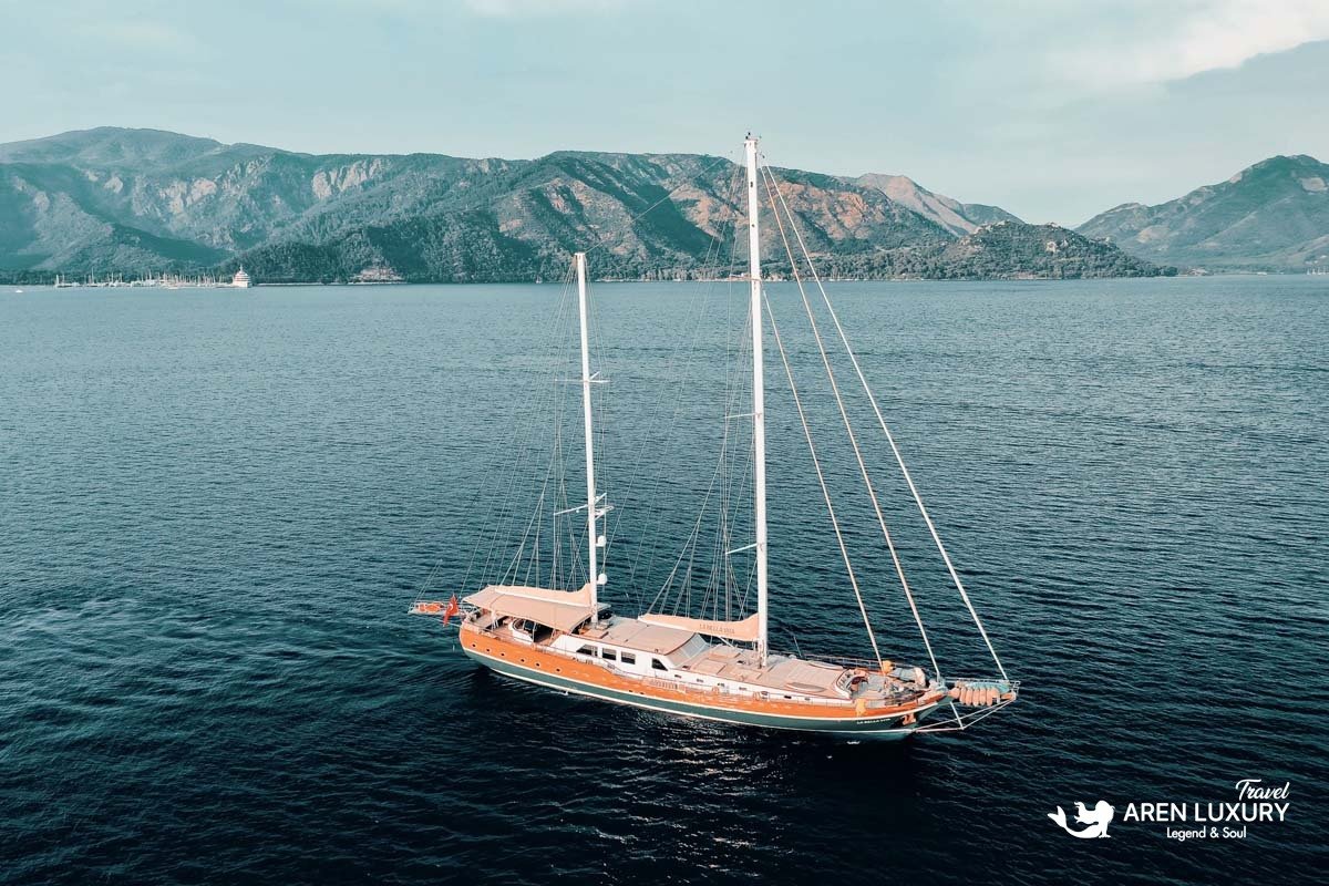 Wide-angle aerial view of the La Bella Vita gulet cruising near a mountainous coastline with its sails lowered.