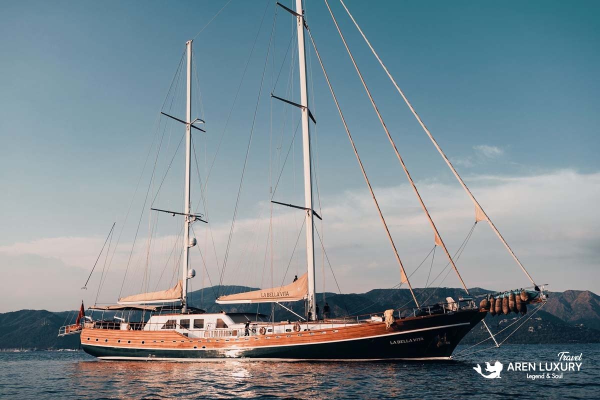 Side profile of the luxury wooden gulet La Bella Vita at anchor on a calm sea with a mountain backdrop.