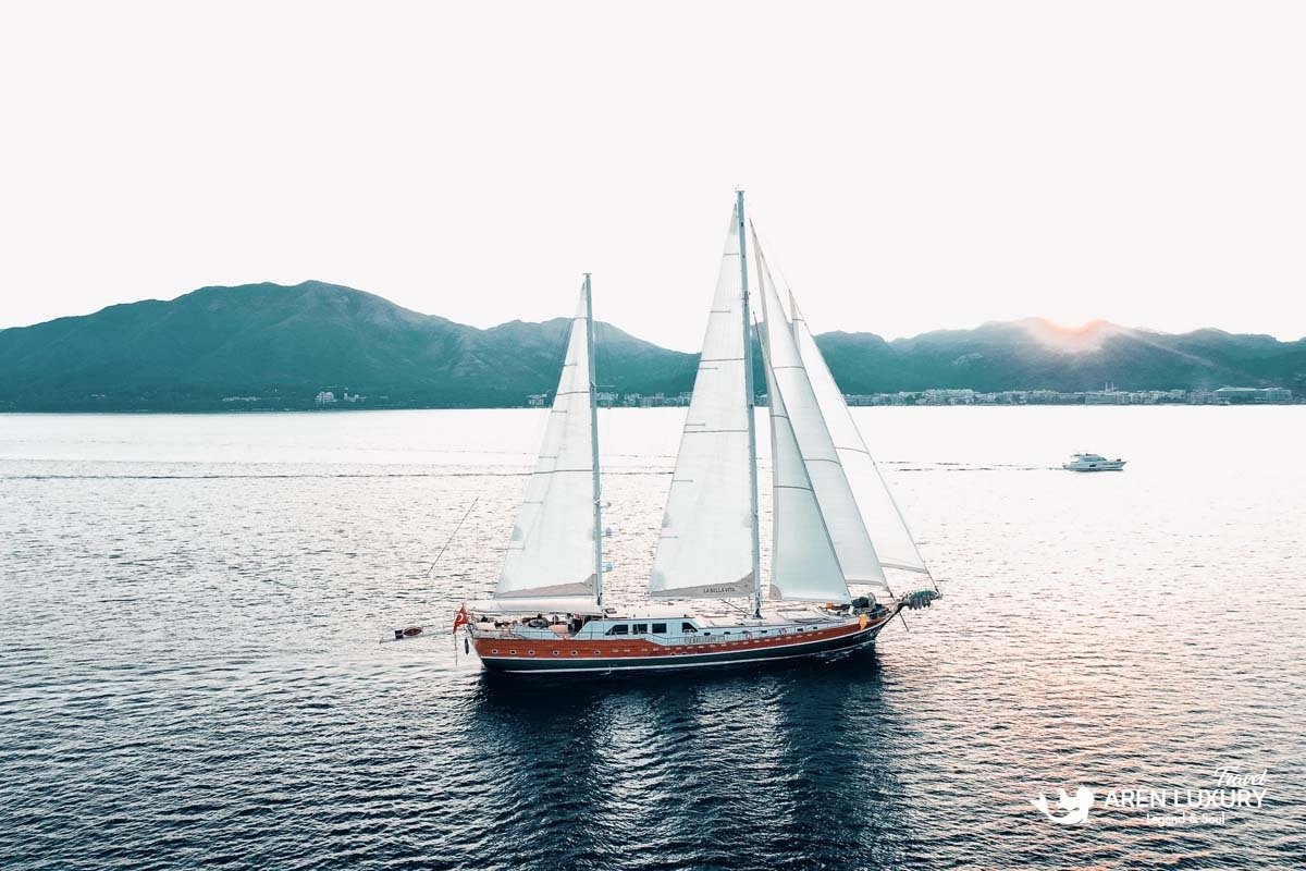 Bright, sunlit wide shot of the La Bella Vita gulet sailing with full white sails on shimmering water with mountains in the background.