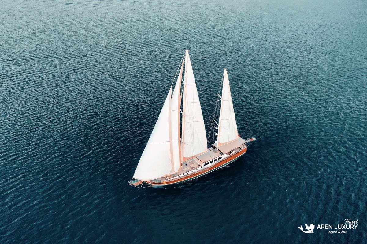 High-angle aerial view of luxury gulet La Bella Vita sailing with full white sails on the deep blue sea.