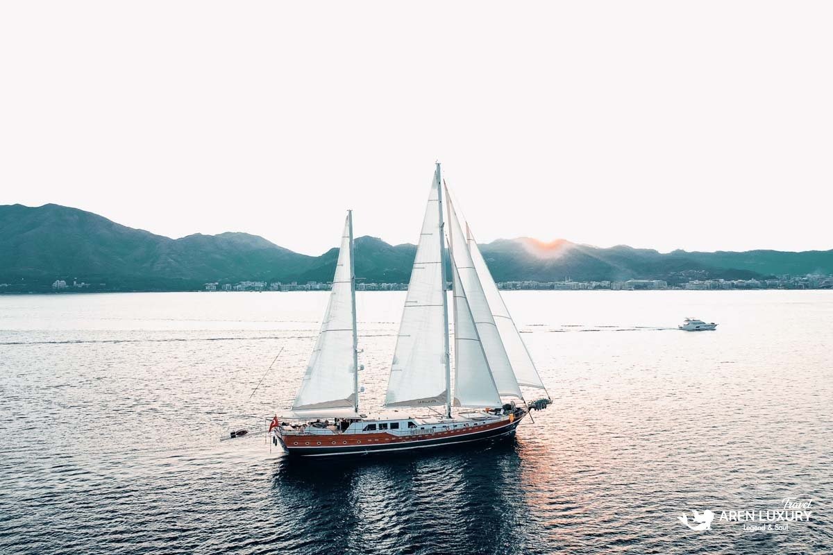 Wide aerial view of the luxury gulet La Bella Vita sailing under full white sails in a calm blue bay surrounded by hills.