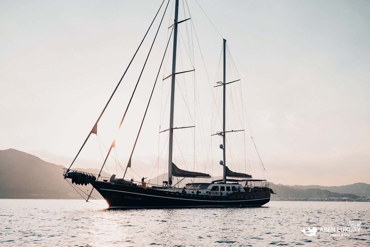 Side profile of luxury wooden gulet La Bella Vita at anchor during sunset with mountains in the background.