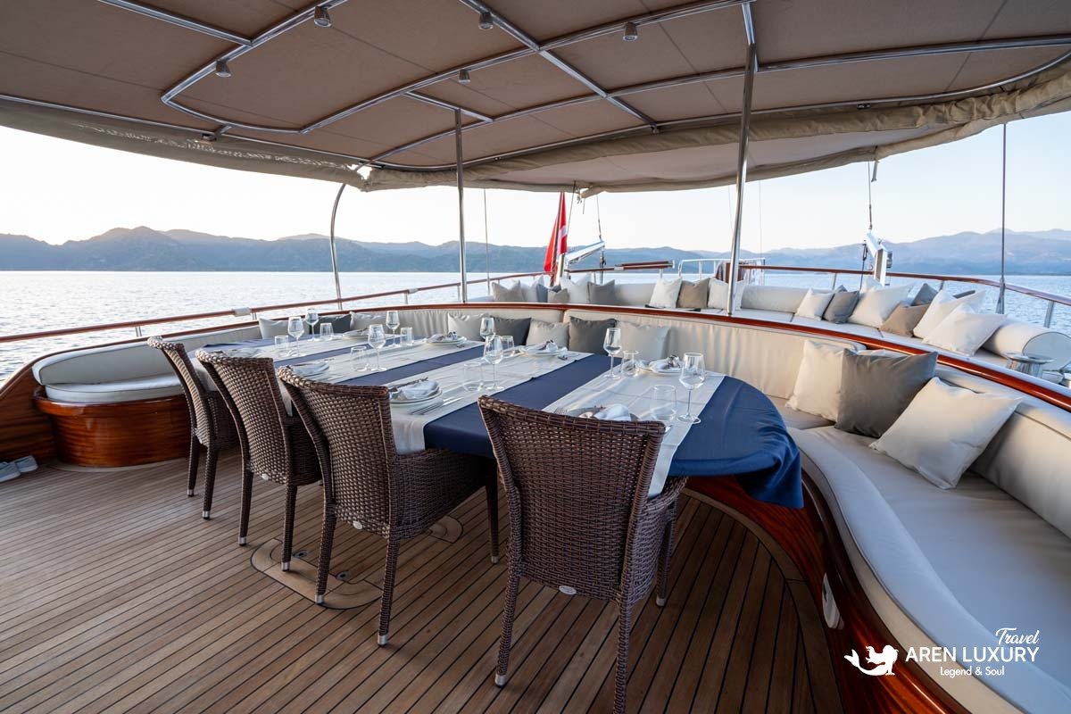 Luxury aft deck on La Bella Vita gulet featuring a large dining table, curved lounge seating, and panoramic sea views.