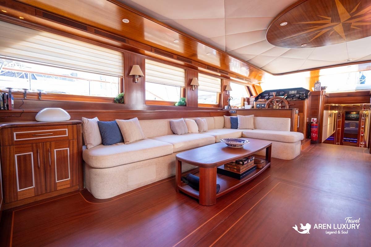 Main salon of a luxury Turkish gulet featuring a cream sectional sofa, polished mahogany wood paneling, and a nautical-themed ceiling.