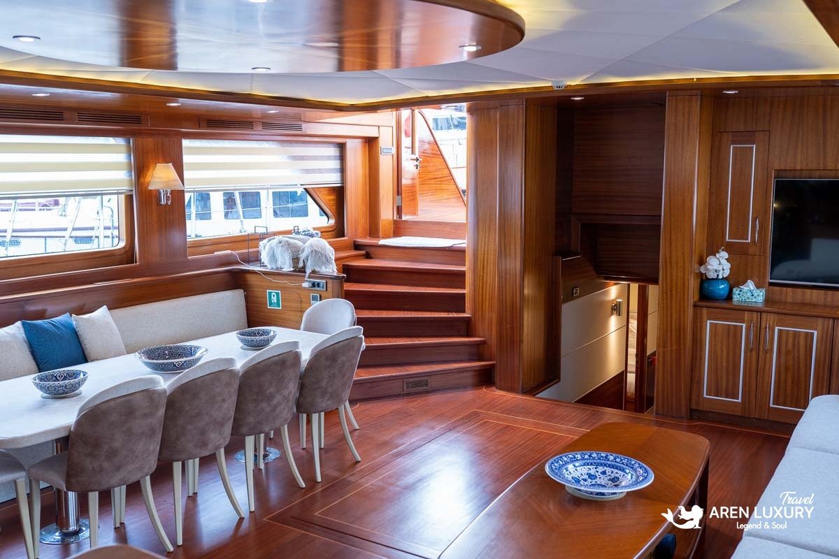 Interior of a luxury yacht main salon showing a large cream sofa, mahogany coffee table, and the captain's wooden steering wheel station.