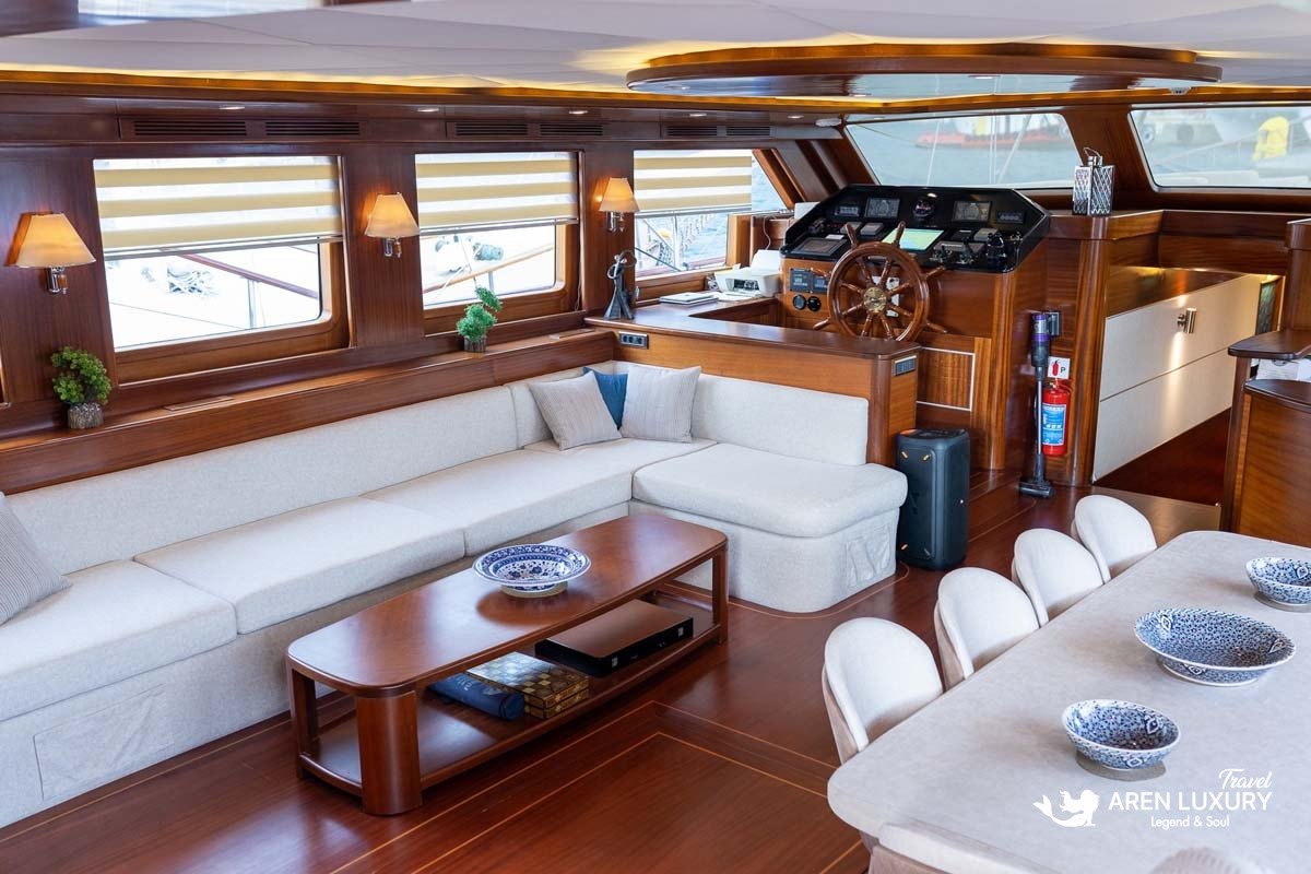 Interior of a luxury yacht main salon featuring a cream sectional sofa, mahogany coffee table, and a classic wooden ship's wheel at the helm station.