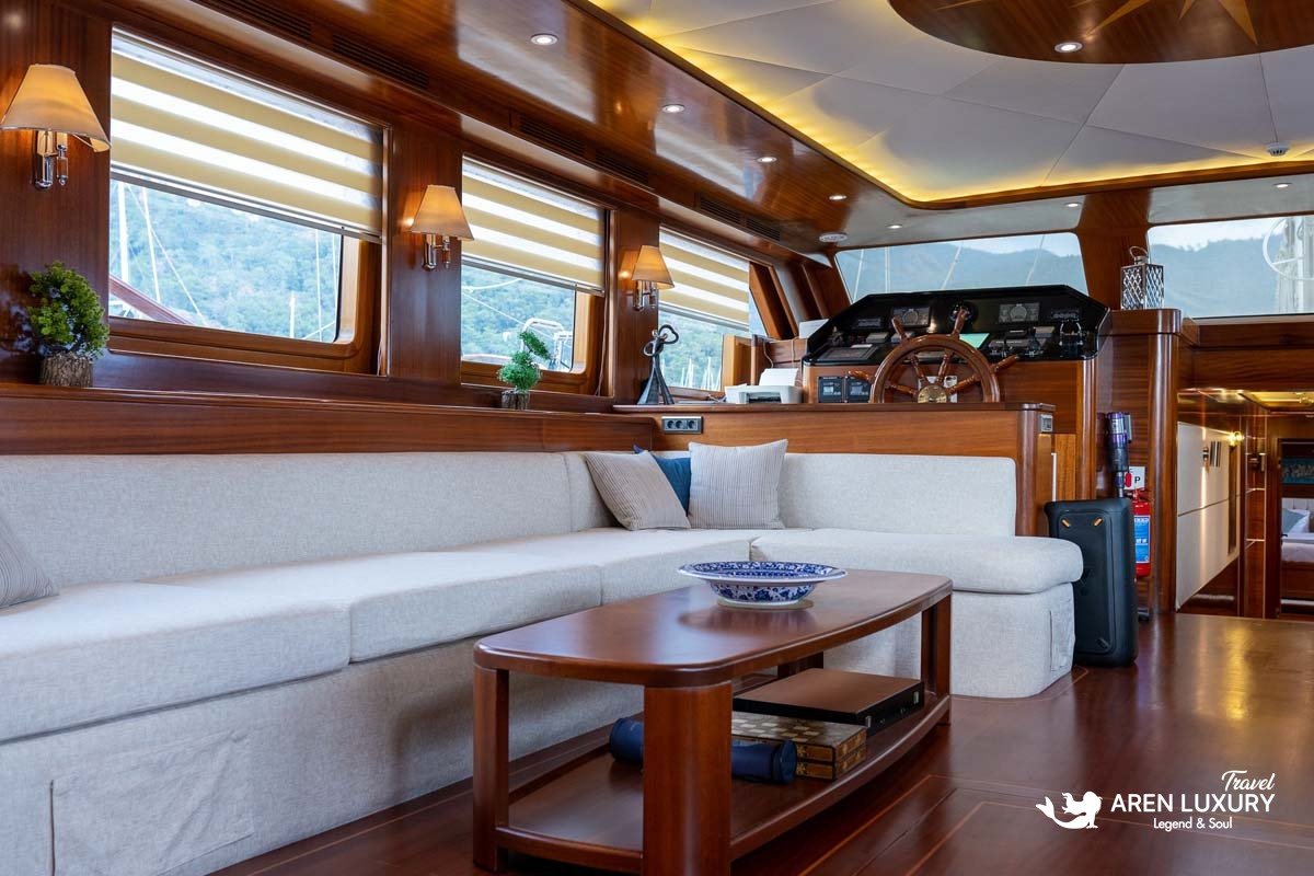 Interior of a luxury yacht main salon featuring a cream sectional sofa, mahogany coffee table, and a classic wooden ship's wheel at the helm station.
