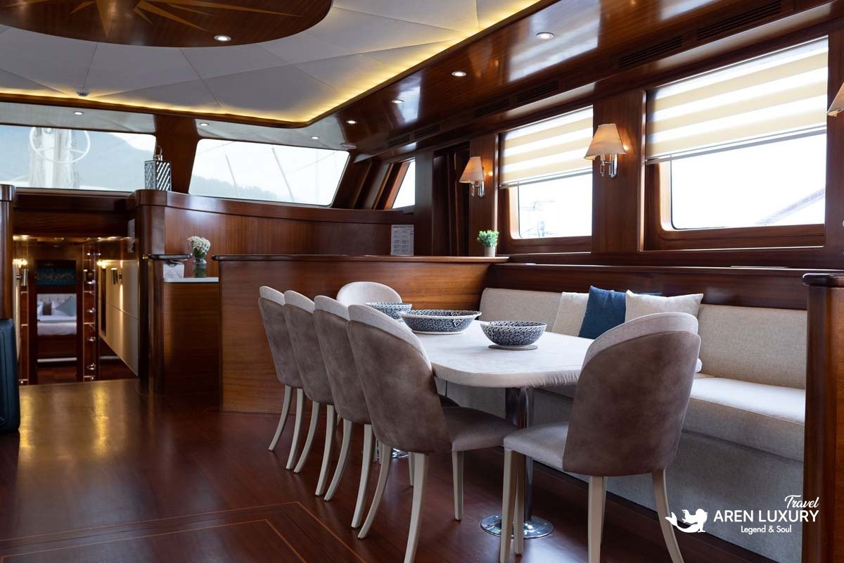 Interior of a luxury yacht main salon featuring a cream sectional sofa, mahogany coffee table, and a classic wooden ship's wheel at the helm station.