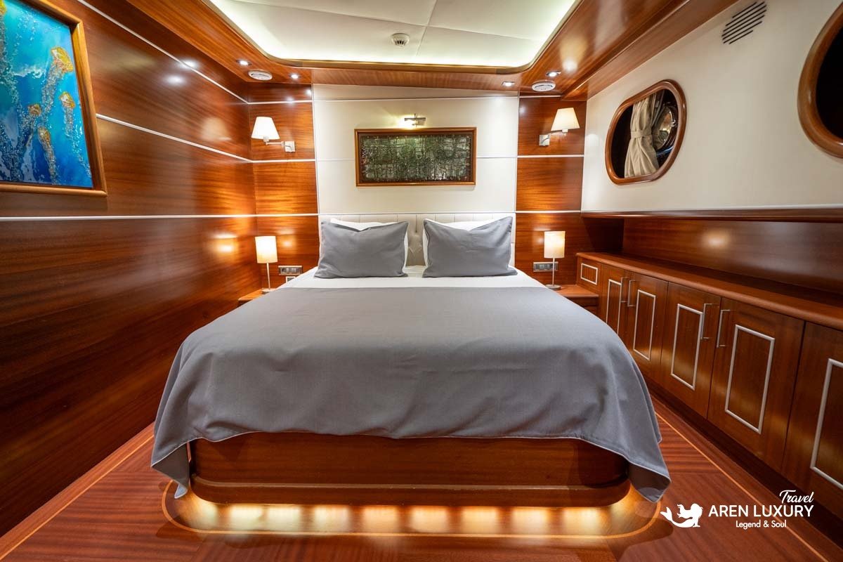 Luxury double guest cabin on La Bella Vita gulet featuring a large bed with warm LED under-lighting, mahogany wood paneling, and nautical porthole windows.