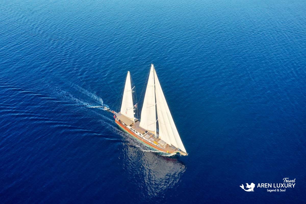Aerial bird's-eye view of luxury gulet La Bella Vita sailing with full white sails on deep blue sea.