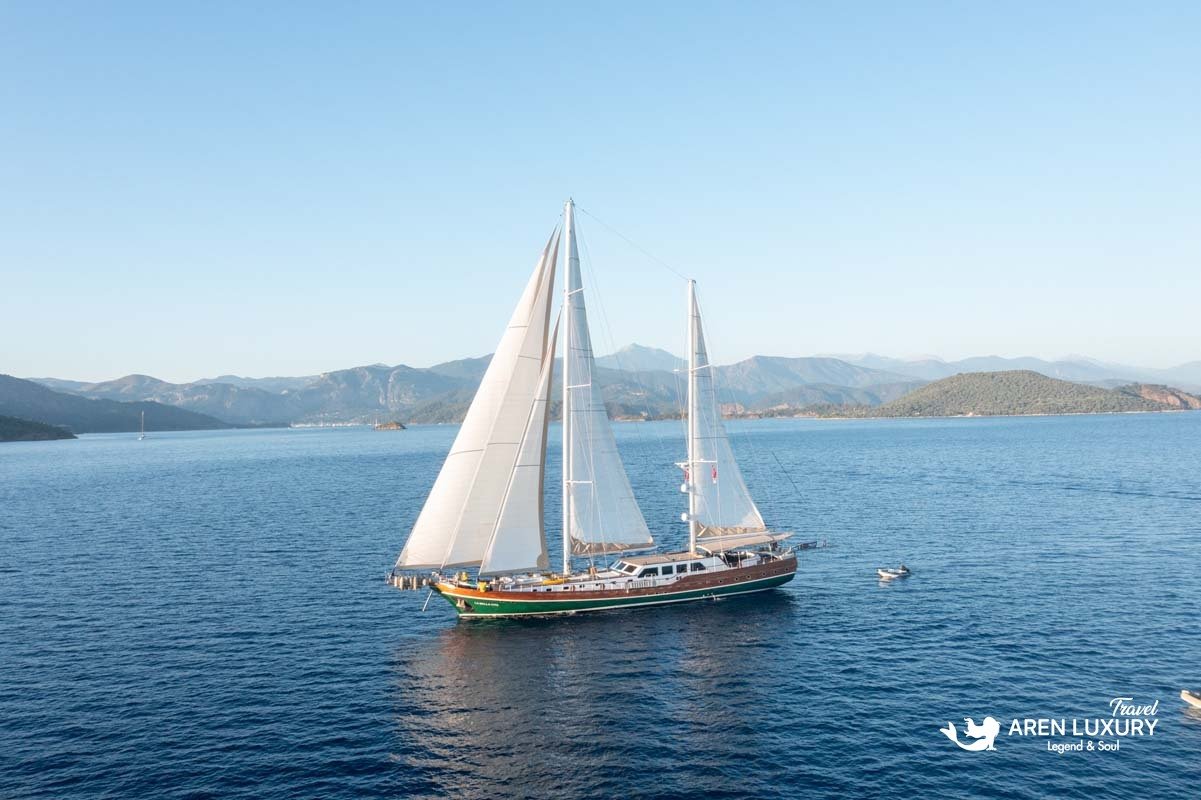 Side profile of La Bella Vita gulet sailing with white sails against a backdrop of green coastal mountains.