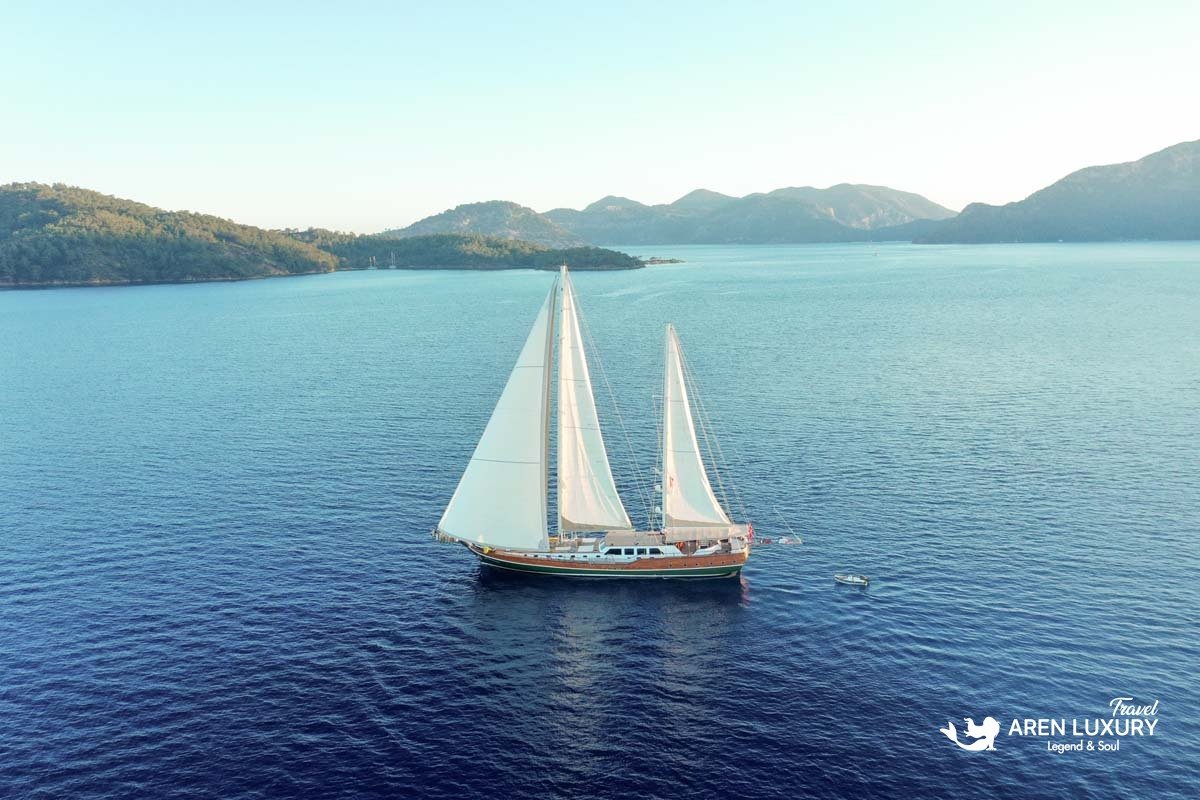Aerial wide shot of the La Bella Vita gulet sailing across a calm blue bay with mountain scenery in the background.