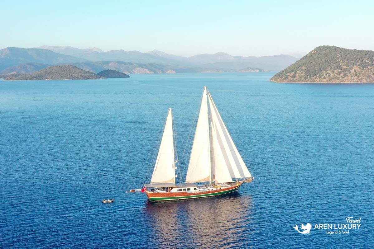 Wide aerial view of the luxury gulet La Bella Vita sailing under full white sails in a calm blue bay surrounded by hills.